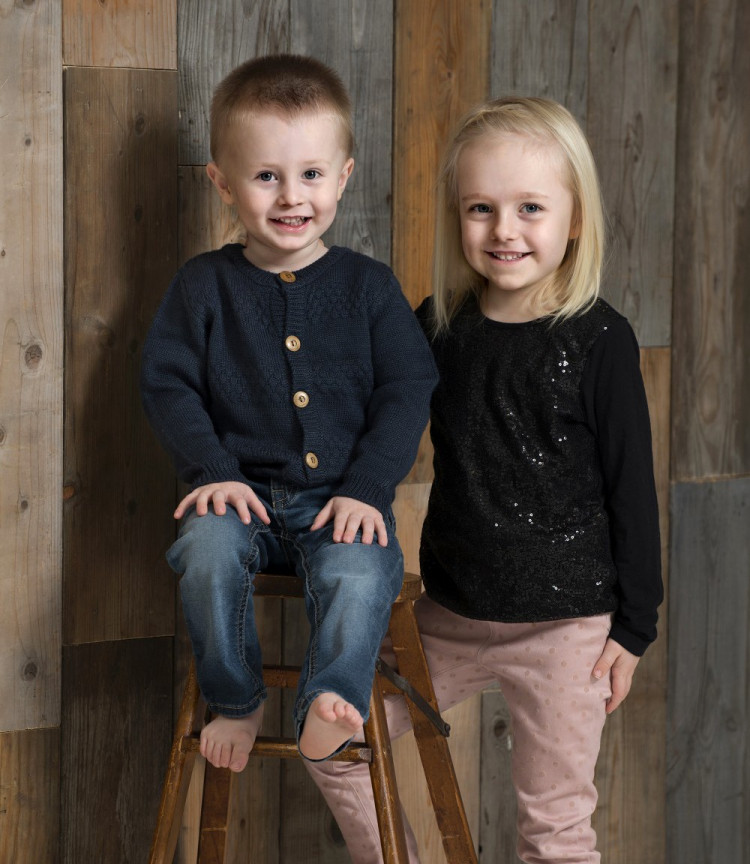 Portrætbillede af to søskende, lillebror og storesøster, med trævæg som baggrund