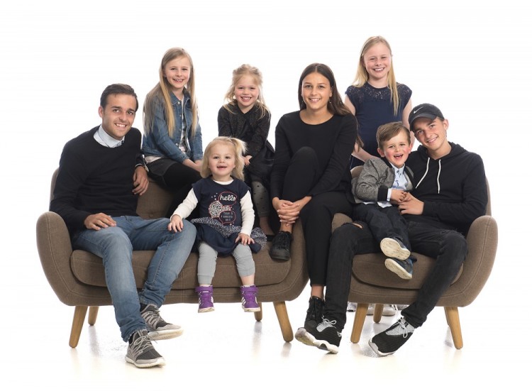 Gruppebillede af otte børnebørn, der sidder og står omkring brun sofa og stol