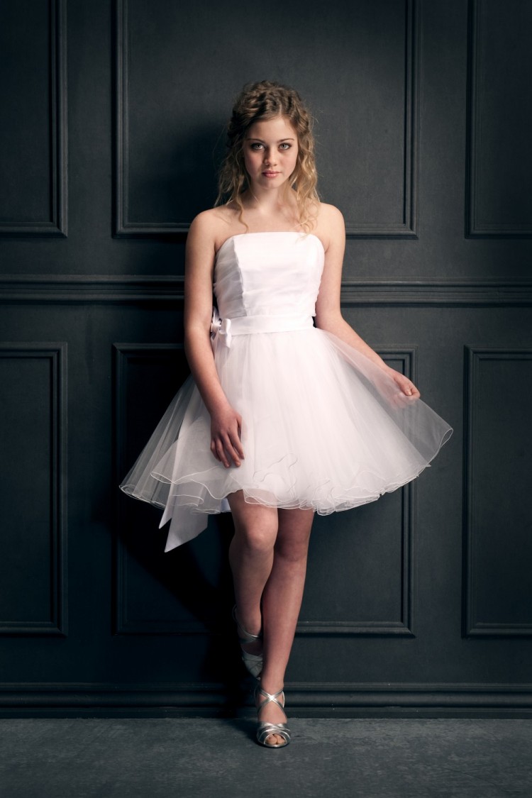 Konfirmationsbillede hvor ung pige i hvid kort kjole står lænet op ad væggen