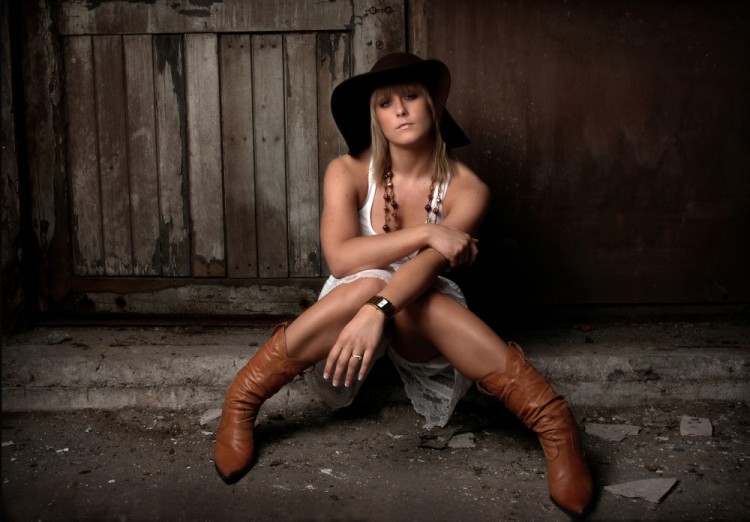 Portræt af ung kvinde med cowboystøvler og cowboyhat, siddende på gulvet i en stald