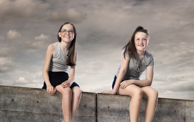 Børnebillede af to piger, der sidder på betonmur med grå himmel som baggrund