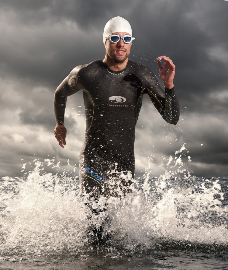 Billede af mand iført våddragt, svømmebriller og badehætte løber i vandet, der sprøjter op omkring ham