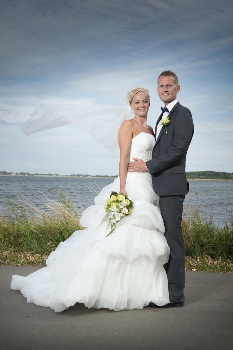 Billede af brudepar, der står foran vand i blæsevejr – sløret blafrer i vinden